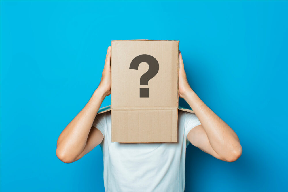 Young man in a white T-shirt with a cardboard box on his head makes a gesture with his hands on a blue background.